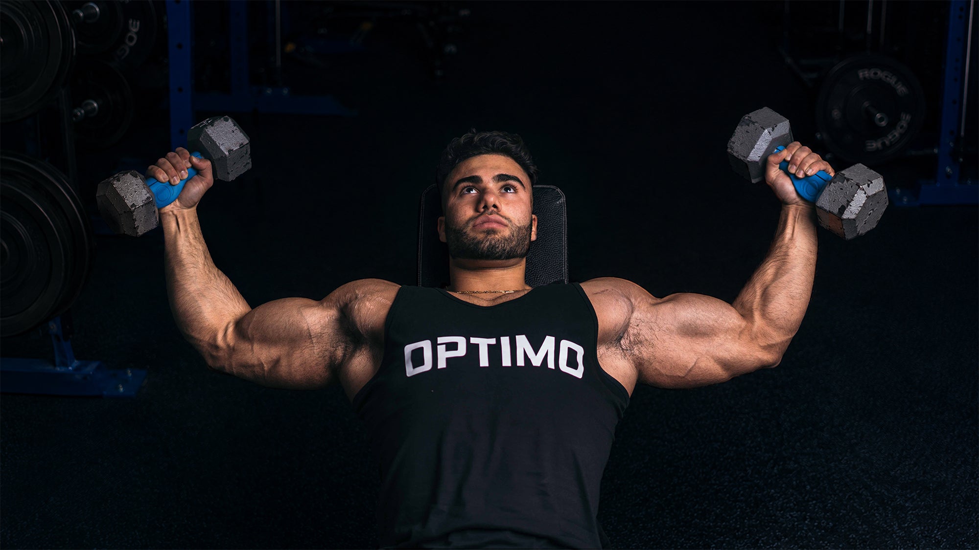 A muscular man lying on a gym bench, holding dumbbells with Optimo One ergonomic thick grips in both hands and wearing a black tank top with the word 'OPTIMO' printed in white across the chest.